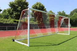 Ein weißes Fußballtor mit rot/weißen Netz und Rädern auf Fußballplatz